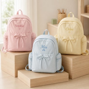 Könnte beinhalten: Drei Rucksäcke in Pastellfarben: ein rosa Rucksack mit der Stickerei 'Sophie' und einem Herz, ein hellblauer mit 'Mia' und einem Regenbogen sowie ein gelber mit 'Chloe' und einem Stern. Jeder Rucksack hat eine Kordelschleife und Seitentaschen.