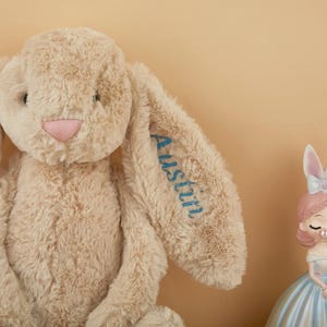 May include: A plush beige bunny with long floppy ears and a pink nose. The name "Austin" is embroidered in blue on one ear. A decorative figurine of a girl in a blue dress and pink hair is also present.