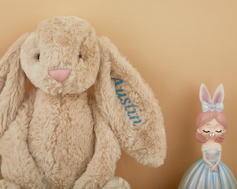 May include: A plush beige bunny with long floppy ears and a pink nose. The name "Austin" is embroidered in blue on one ear. A decorative figurine of a girl in a blue dress and pink hair is also present.