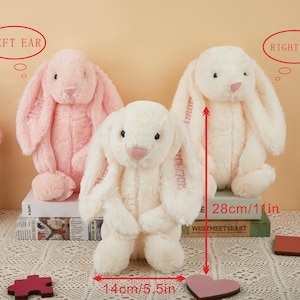 May include: Three plush bunny rabbits in pink and cream colors. The cream-colored bunny is 11 inches tall and 5.5 inches wide. The bunnies have long floppy ears and embroidered details. The bunnies are sitting on books and a lace surface.