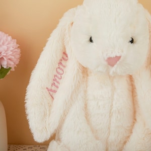 May include: A plush, cream-colored bunny with long, floppy ears. The bunny has a pink nose and the word "Amoru" embroidered in pink on one ear. The soft toy is set against a light yellow background, with a vase of pink flowers visible on the left.