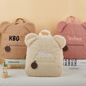 May include: Three teddy bear-shaped backpacks in tan, cream, and pink. Each backpack has a zippered pocket, a top handle, and embroidered text. The tan backpack has "KBG" embroidered, the cream one has "Mama", and the pink one has "Vedina".