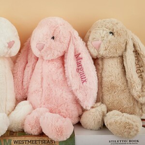 May include: Three plush bunny rabbits in cream, pink, and tan. The pink bunny has the name "Margaux" embroidered on its ear. The tan bunny has the name "Freddv" embroidered on its ear. The bunnies have long floppy ears and soft, rounded bodies.