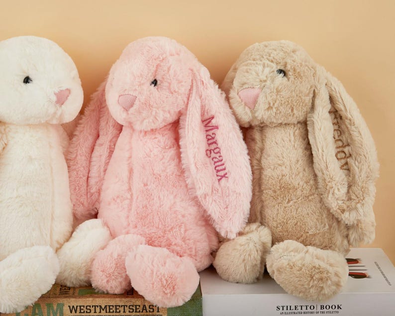 May include: Three plush bunny rabbits in cream, pink, and tan. The pink bunny has the name "Margaux" embroidered on its ear. The tan bunny has the name "Freddv" embroidered on its ear. The bunnies have long floppy ears and soft, rounded bodies.