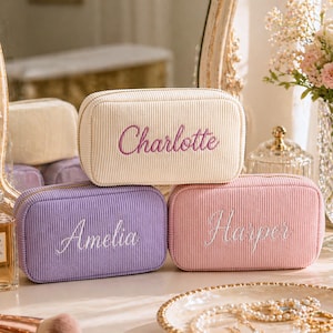 Op de afbeelding: Drie corduroy make-uptassen in crème, lavendel en roze, elk geborduurd met een naam in een complementaire kleur. De crèmekleurige tas zegt "Charlotte" in paars, de lavendelkleurige tas zegt "Amelia" in wit en de roze tas zegt "Harper" in wit.