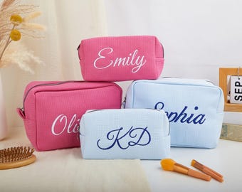 May include: Four personalised cosmetic bags in pink and blue. Each bag has a different name or initials embroidered in a contrasting colour. The bags are rectangular with a zip closure, ideal for storing makeup and toiletries.