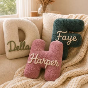 May include: Three letter-shaped throw pillows made of textured cream, dark green, and dusty rose fabric. The cream pillow features the name Della in green script, the green pillow features Faye in white script, and the rose pillow features Harper in white.