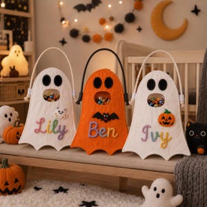 May include: Three felt ghost-shaped treat bags with rope handles. Two are white and one is orange. Each features a cutout mouth filled with colorful candies, black felt eyes, and embroidered names: Lily, Ben, and Ivy. Decorative felt appliques include a cat, a bat, and a pumpkin.