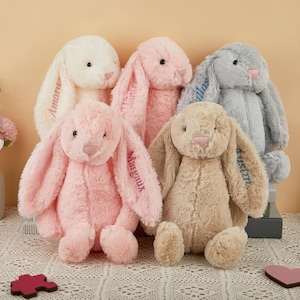 May include: Five plush bunny rabbits in cream, pink, and gray hues. Each bunny has long, floppy ears and embroidered names. The bunnies are soft and cuddly, perfect for children. The bunnies are sitting on a white lace tablecloth.