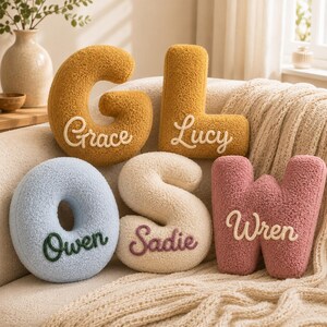 May include: Five plush letter-shaped cushions with a textured, sherpa-like finish. The mustard yellow G features the name Grace, the mustard yellow L features Lucy, the light blue O features Owen, the cream S features Sadie, and the pink W features Wren.