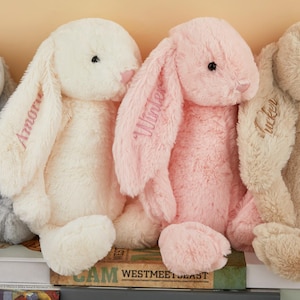 May include: A row of plush bunny rabbits in various pastel colors, including gray, cream, and pink. Each bunny has long, floppy ears with embroidered names. The bunnies are soft and cuddly, perfect for children.
