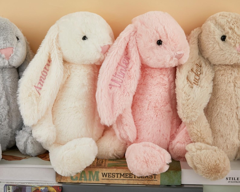 May include: A row of plush bunny rabbits in various pastel colors, including gray, cream, and pink. Each bunny has long, floppy ears with embroidered names. The bunnies are soft and cuddly, perfect for children.
