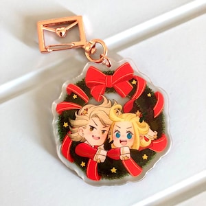 May include: A Christmas-themed acrylic keychain featuring two cartoon characters, a blonde male and a blonde female, embracing inside a green wreath with red ribbon and gold stars.