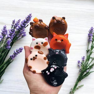 Könnte beinhalten: Eine Sammlung von sieben kleinen, runden, tierförmigen Keramikschalen in verschiedenen Farben wie Braun, Weiß, Orange, Schwarz und Hellrosa. Jede Schale zeigt ein stilisiertes Tiergesicht mit Ohren und Pfoten, gestaltet als kleine Ablage oder Stäbchenbank.