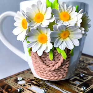 Könnte beinhalten: Eine weiße Keramiktasse mit einem 3D-Design eines Korbs mit weißen Gänseblümchen mit gelben Zentren und grünen Blättern. Die Gänseblümchen sind aus Polymer Clay gefertigt und der Korb hat ein geflochtenes Design.