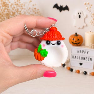 May include: A white ghost keychain featuring a textured orange knit-style hat, black eyes, a small smile, and pink cheeks. The ghost is holding a small orange pumpkin with green leaves. The keychain includes a silver metal ring and chain attachment.