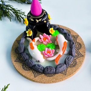 Op de afbeelding: Een decoratieve wierookbrander met een miniatuur koi-vijver scène. De vijver bevat twee wit-oranje koi-vissen, roze lotusbloemen en groene waterleliebladeren. De brander is zwart met gouden accenten en een felroze wierookkegel.