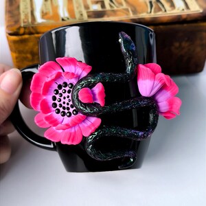 May include: Black ceramic mug with a pink flower and a black snake with iridescent green accents. The snake is wrapped around the flower.