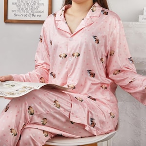 Op de afbeelding: Roze pyjamaset met een patroon van honden en pootafdrukken. De top heeft lange mouwen, een knoopsluiting en een kraag. De bijpassende broek maakt de set compleet. De pyjama is gemaakt van een zacht, zijdeachtig materiaal. De tekst op de muur luidt "You're still the one".
