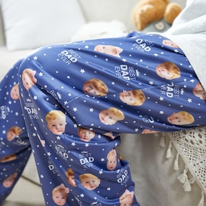 Puede incluir: Pantalones de pijama azules con un estampado de caras de bebé, estrellas blancas y el texto "BEST DAD EVER". Los pantalones están hechos de un material suave y sedoso.