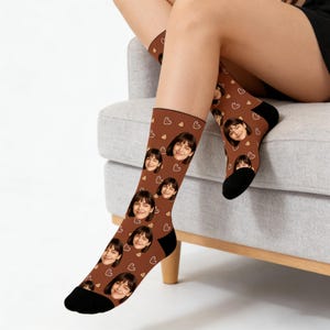 May include: Brown socks featuring a repeating pattern of a smiling face and small white hearts. The socks have black heels and toes. The socks are worn by a person sitting on a grey couch.