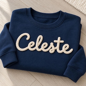 May include: A navy blue long-sleeve sweatshirt featuring the name Celeste written in a raised, cream-colored rope-style script across the chest. The garment has a ribbed crew neckline and ribbed cuffs on the long sleeves.