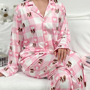 Puede incluir: Conjunto de pijama rosa y blanco con estampado a cuadros y estampado de cara de perro. La pijama tiene mangas largas y pantalones largos.
