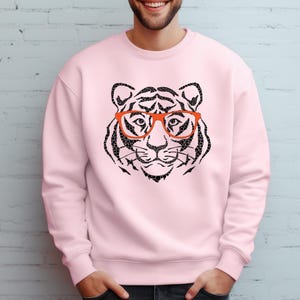 May include: A pink sweatshirt featuring a black and white tiger wearing orange glasses. The tiger is a graphic print on the front of the sweatshirt.