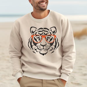 May include: A beige sweatshirt featuring a black and white graphic of a tiger wearing orange glasses.