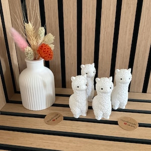 Könnte beinhalten: Vier weiße Keramik-Alpaka-Figuren auf einem Holzregal mit einer weißen Vase und getrockneten Blumen. Das Regal hat schwarze Linien, die vertikal verlaufen. Die Figuren sind alle nach vorne gerichtet und haben schwarze Augen. Die Vase ist gerippt und hat einen weißen Hintergrund. Die Blumen sind getrocknet und haben eine rosa und orange Farbe.