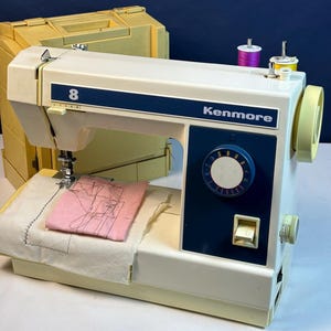 May include: A vintage Kenmore sewing machine, model 8, in cream and navy blue. The machine has a pink fabric square with a stitched design, and two spools of thread. A yellow carrying case is in the background.