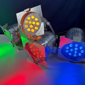 May include: Several stage lights with different colored LED lights. One light emits green light, another red, another blue, and another yellow. The lights are in a silver metal housing and are angled towards the viewer.