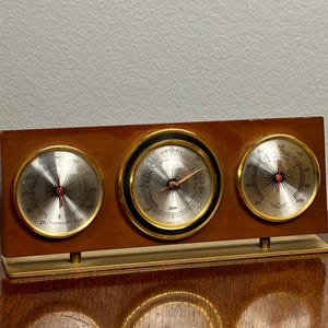 May include: A rectangular wooden weather station featuring three circular gauges with gold-toned rims. The left gauge displays temperature in Fahrenheit, the center barometer shows weather conditions, and the right gauge indicates relative humidity.