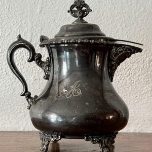 May include: Antique silver-plated creamer with a lid and ornate handle. The creamer features a decorative spout and is supported by three ornate feet. An engraved initial "A" is visible on the body of the creamer.