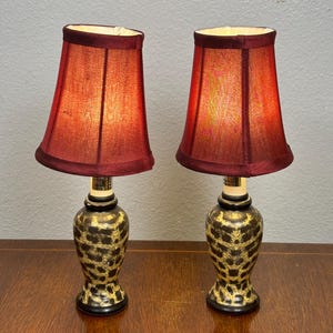 May include: A pair of table lamps featuring dark red fabric shades and bulbous bases with a black and gold leopard print pattern. Each lamp has a narrow neck and a flared base, providing a consistent look for both lighting fixtures.