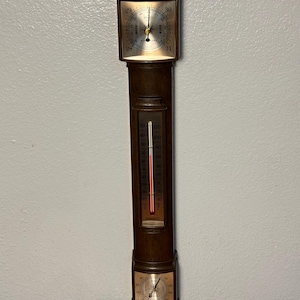 May include: A vintage wooden barometer with a square top featuring a dial indicating pressure readings. Below, a thermometer displays temperature in Fahrenheit and Celsius. The bottom section has a circular dial. The device is mounted on a wall.