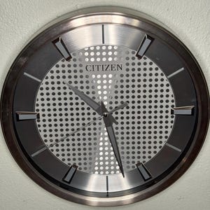 May include: A round Citizen wall clock with a silver-tone metal frame. The clock face has a perforated silver center and a dark gray outer ring. The hands and hour markers are dark gray. The word "CITIZEN" is printed at the top of the clock face.
