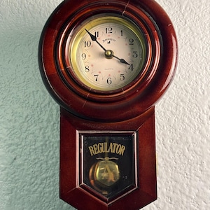 May include: A vintage Remington quartz wall clock with a dark brown wooden frame. The clock face is cream-colored with black numerals and hands. The lower section features a glass panel with the word "REGULATOR" and a pendulum.