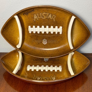 May include: Two brown football-shaped chip bowls. Each bowl has white accents and a textured surface. The words "Official", "Allstar", and "Chip Bowl" are printed on the bowls. The bowls are stacked on top of each other.
