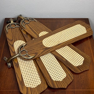 May include: A set of three ceiling fan blades featuring a dark brown wood grain finish with a central inset of light beige woven cane webbing. Each blade is attached to a decorative antique brass metal mounting bracket with a curved design.