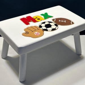 May include: A white wooden step stool with the name "Max" in red, yellow, and green letters. The stool is decorated with a baseball glove, soccer ball, and football. The stool has four white legs.