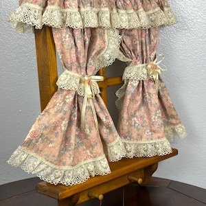 May include: A miniature wooden window frame with pink floral curtains and a matching valance. The curtains are trimmed with cream-colored lace and tied back with ribbons and small pink roses. The window frame has a small shelf with pegs.