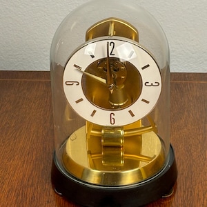 May include: A gold-toned mantel clock with a white clock face and black numerals, encased in a clear glass dome. The clock has a black base and is set on a wooden surface. The clock's hands point to 12 and 6.