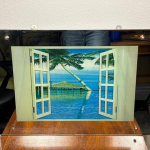 May include: A framed artwork depicting an open window overlooking a tropical scene. The window is white with multiple panes, revealing a palm tree, blue water, and a small structure. The frame is dark with a gold trim.
