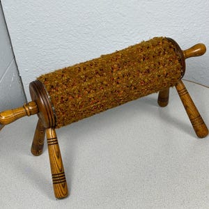 May include: A vintage footstool with a cylindrical, textured, brown and orange fabric seat. The seat is supported by a wooden frame with four legs and two handles. The wooden legs have decorative grooves.