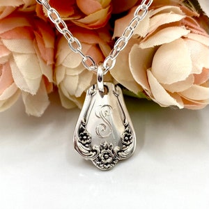 May include: A silver pendant necklace featuring a spoon-shaped charm. The charm has floral detailing and an engraved letter "S". The necklace is suspended from a silver chain, with a soft floral background.