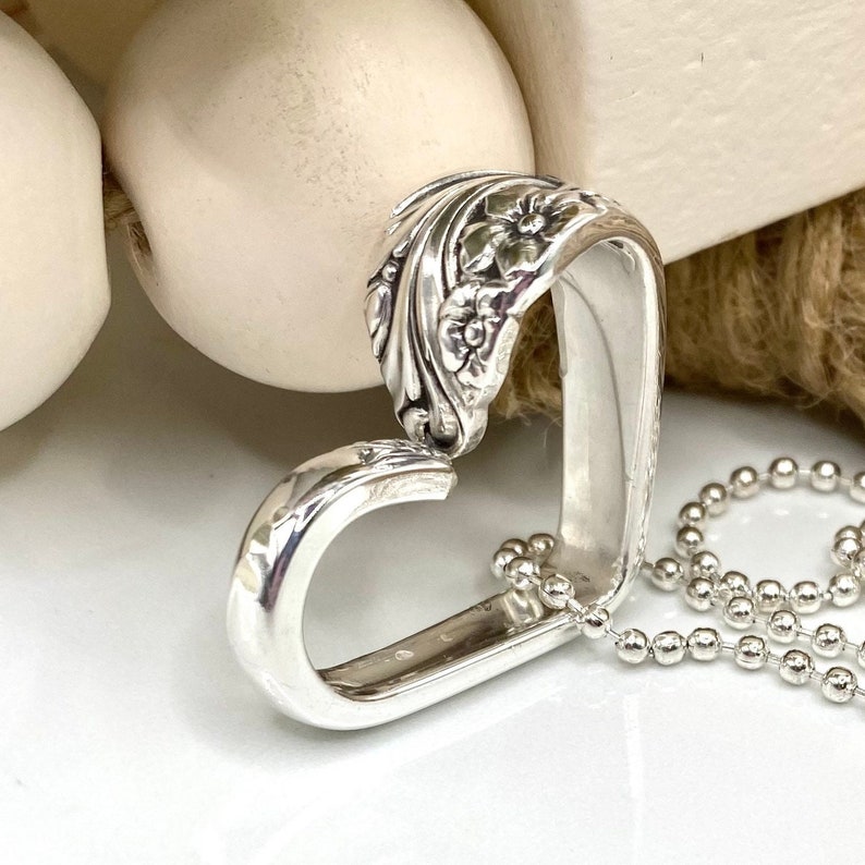 May include: A silver heart-shaped pendant necklace. The pendant is crafted from a spoon, with floral detailing on the top portion. The necklace features a delicate ball chain. The pendant is approximately 1.5 inches in length.