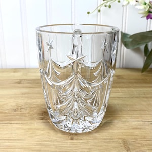 May include: A clear glass mug featuring a molded design of stylized evergreen trees and stars. The transparent vessel has a smooth rim, a sturdy handle, and a textured exterior surface that catches the light, creating a reflective appearance.