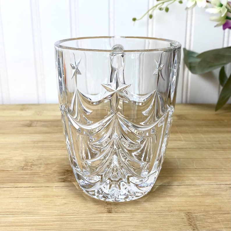 May include: A clear glass mug featuring a molded design of stylized evergreen trees and stars. The transparent vessel has a smooth rim, a sturdy handle, and a textured exterior surface that catches the light, creating a reflective appearance.