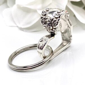 May include: A handcrafted silver key ring made from a repurposed spoon. The spoon's handle is bent to form a loop, and the spoon's bowl is decorated with floral patterns. A silver key ring is attached to the spoon.